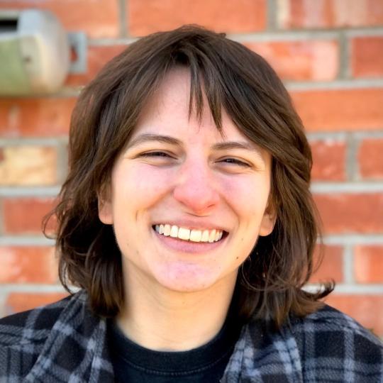 Photo of author Lee Bishop. The are smiling at the camera, with a red brick wall in the background. They have shoulder-length dark brown hair and wear a black t-shirt with a black and blue plaid jacket.