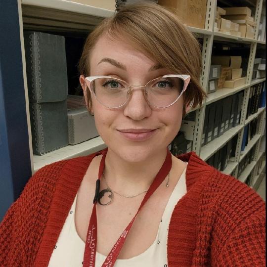 Photo of Cassidy Nemick, who is wearing a white scoop neck shirt underneath a dark orange waffle-knit cardigan. A red lanyard is around their neck, along with a circular pendant of the moon and one star. They have light brown short hair that is parted on their right side, and metal-framed cateye spectacles. The photo is a selfie, and they are looking at the camera and smiling.