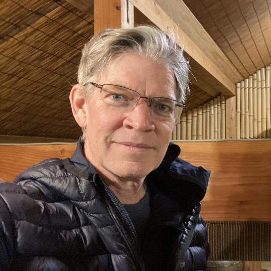 Photo of a middle-aged Caucasian man wearing eyeglasses. He has on a black shirt and a black puffy jacket. He has short-trimmed salt-and-pepper colored hair.