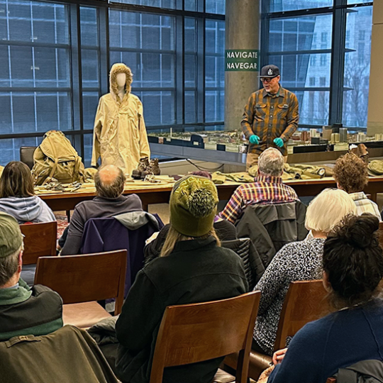A large group of visitors in a rows of chairs sit attentively while a presenter shares numerous artifacts, including many uniforms from the 10th Mountain Division.