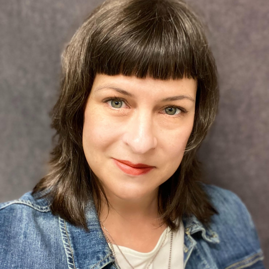 Close-up photo of author Ann Sneesby-Koch. She has shoulder length dark hair with bangs, and she's wearing a denim top. She's smiling at the camera.